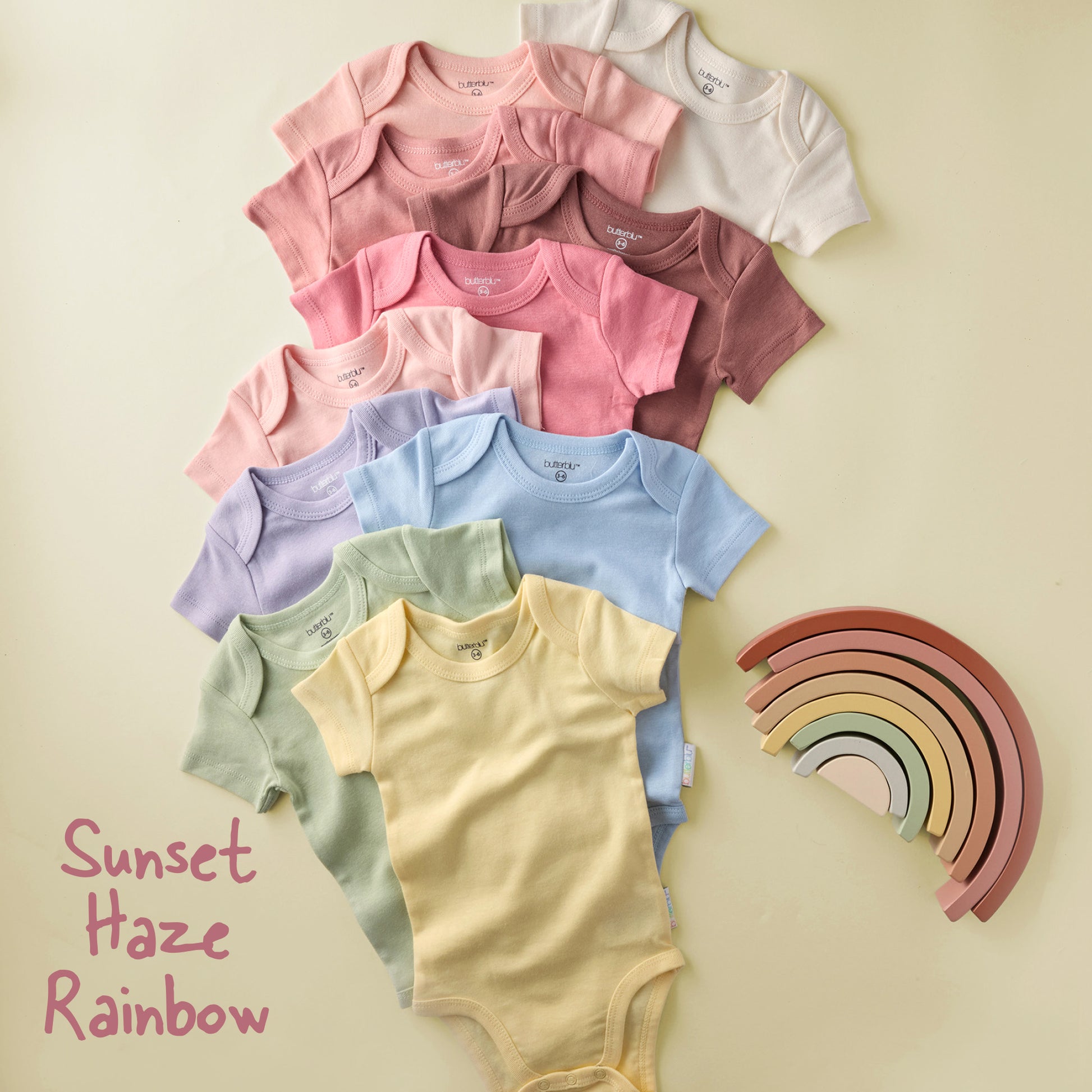 Stack of colorful baby bodysuits with a wooden rainbow toy on a beige background.