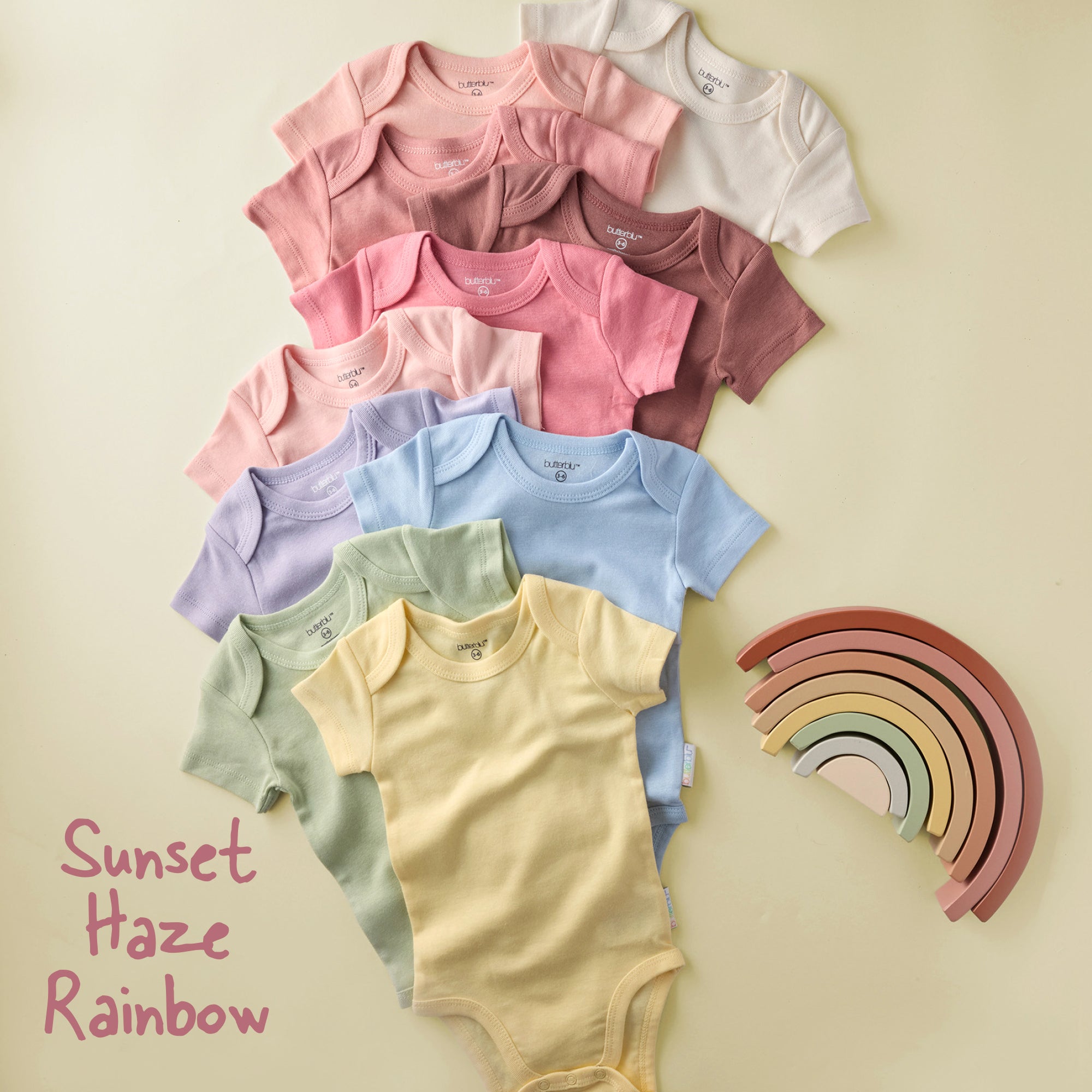 Stack of colorful baby bodysuits with a wooden rainbow toy on a beige background.