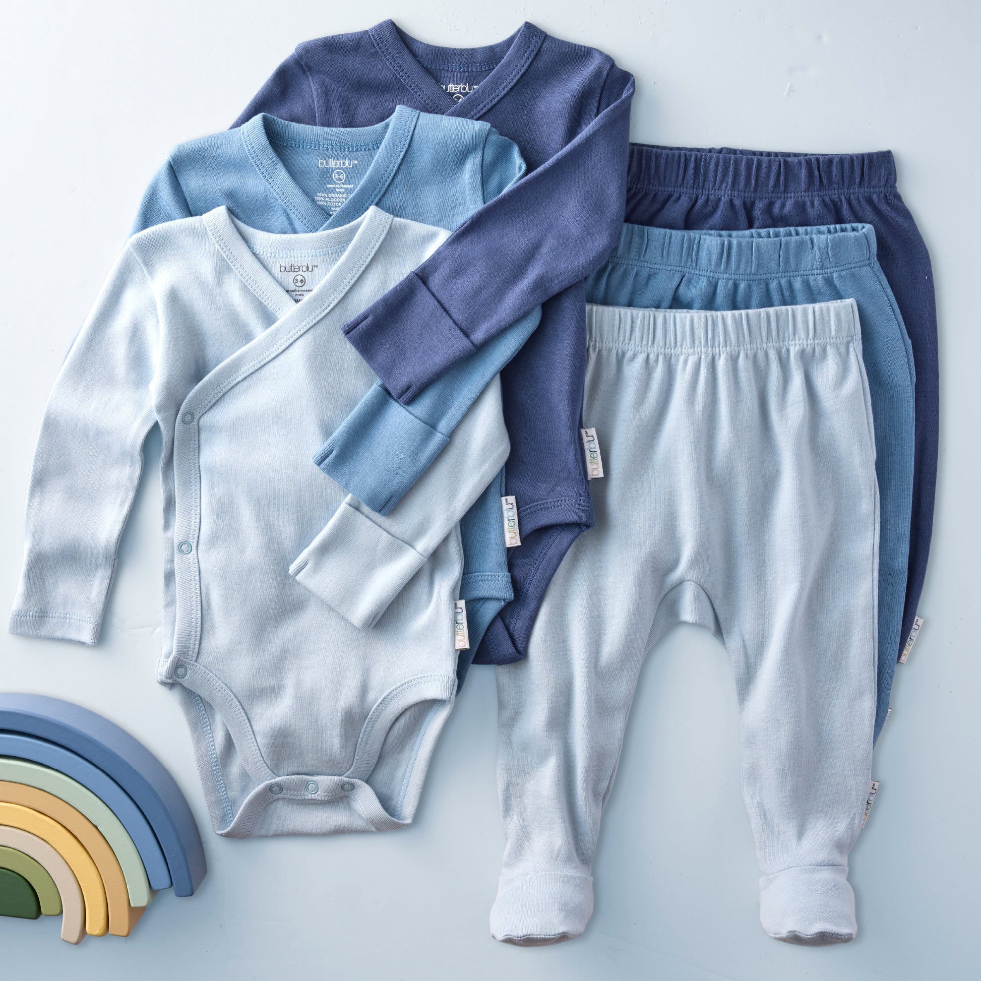 Baby clothing set including a cardigan, onesie, and pants on a light blue background with a wooden rainbow toy.