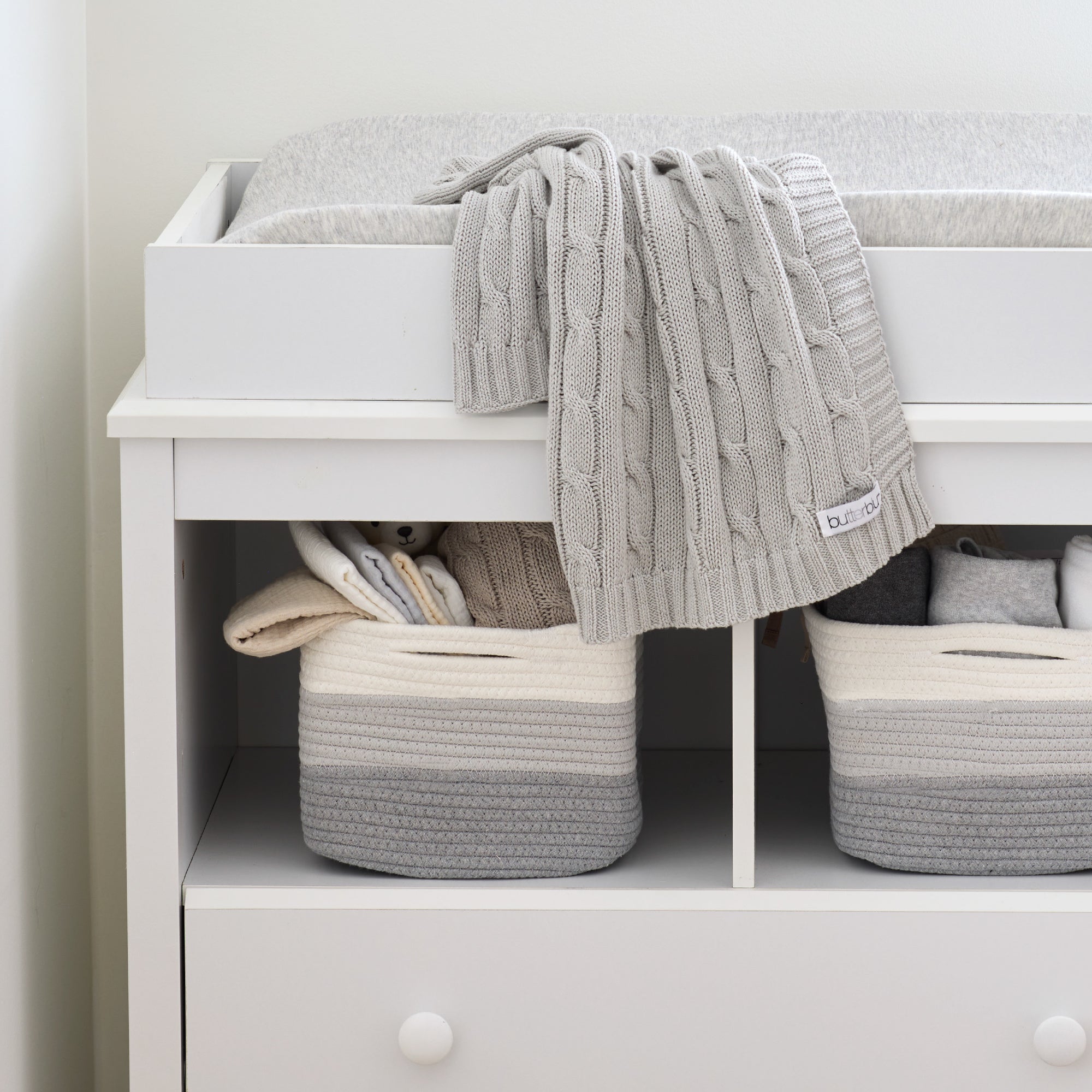 White dresser with gray and beige woven baskets and a gray knit blanket.