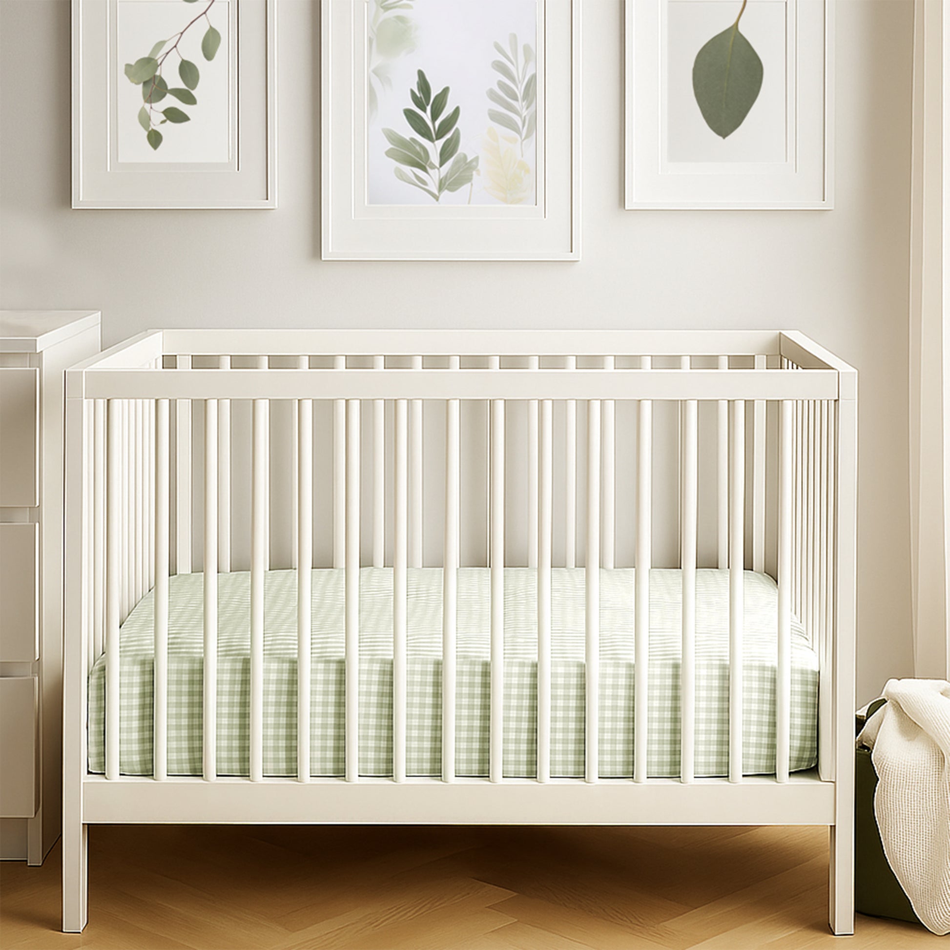 White crib with green bedding in a room with leaf-framed pictures on the wall.