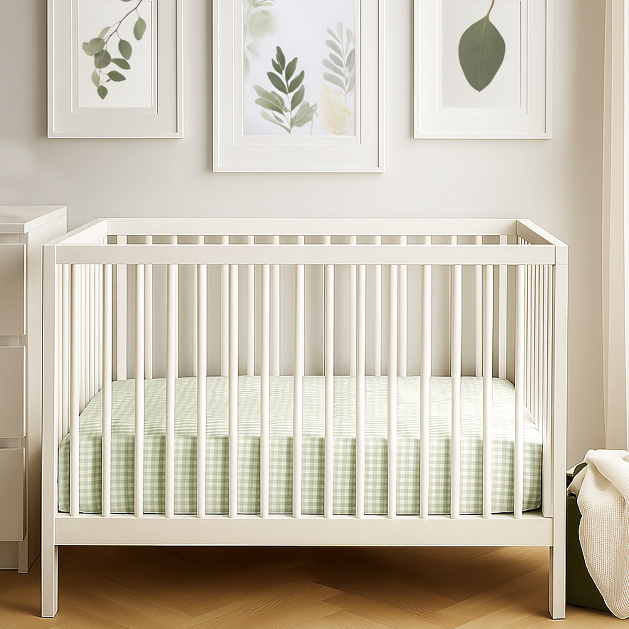White crib with green bedding in a room with leaf-framed pictures on the wall.