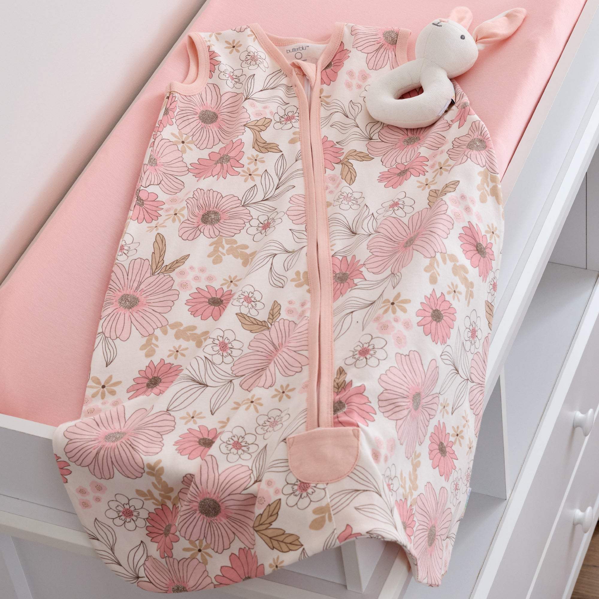 Floral-patterned baby sleeping bag with a white bunny plush toy on a pink surface.
