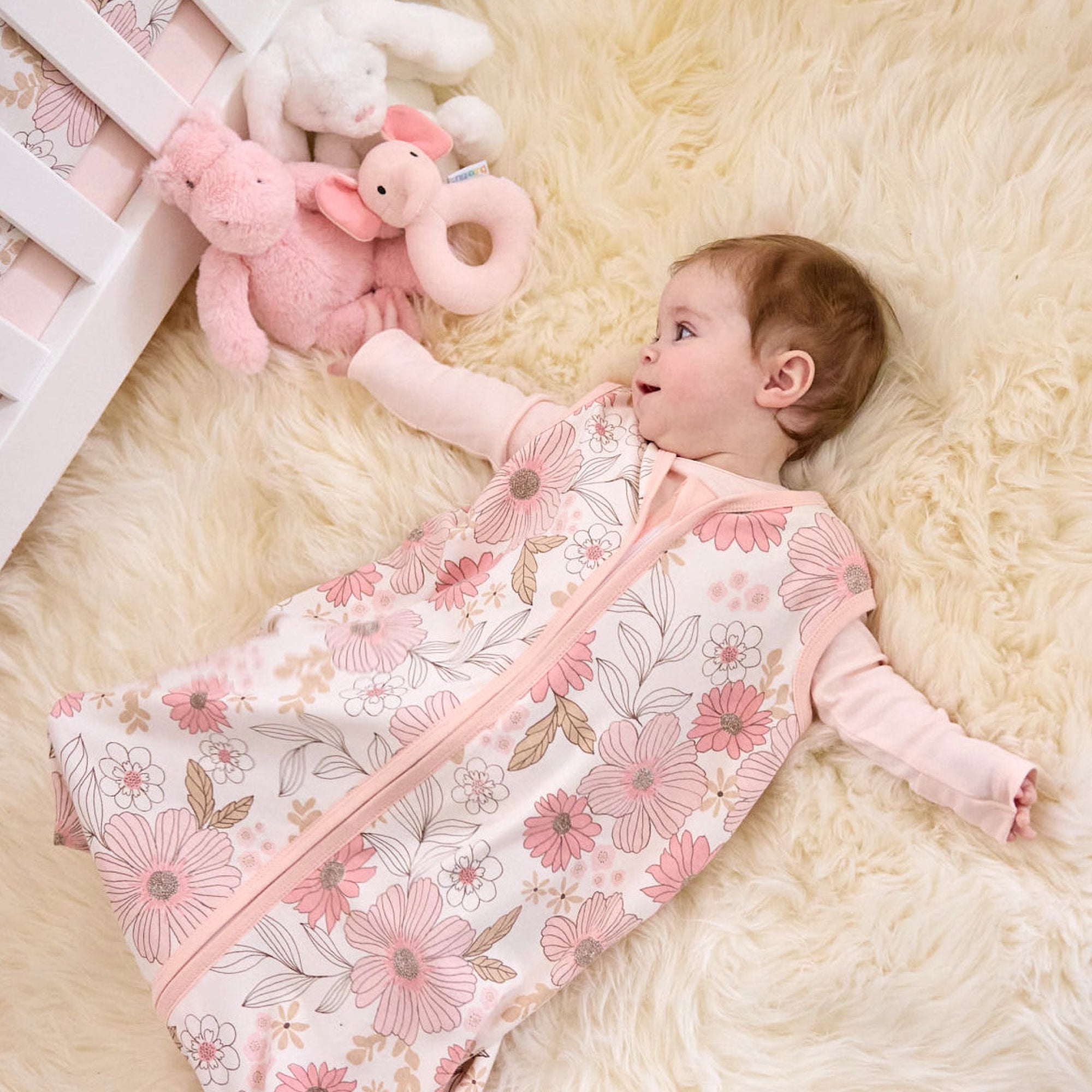 Baby lying on a fluffy white rug with a floral-patterned swaddle and pink stuffed animals.