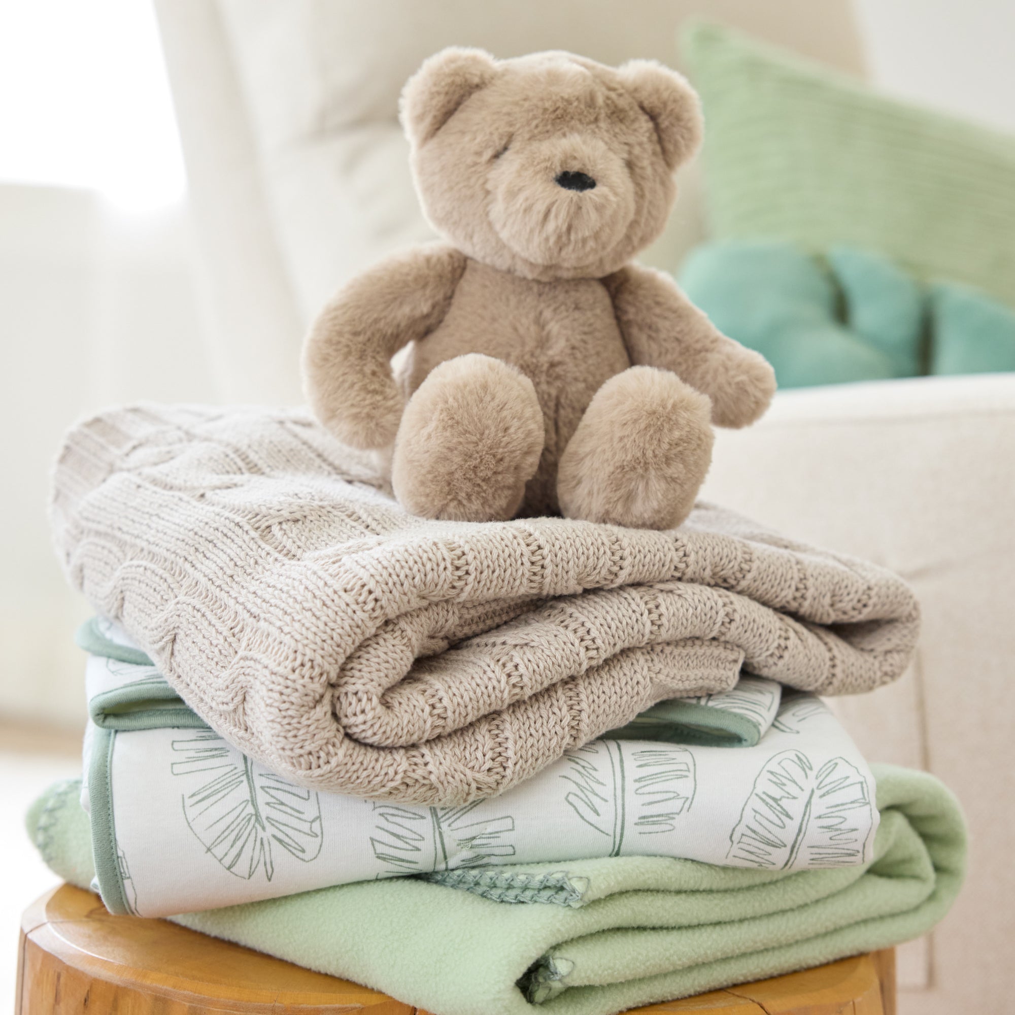 Stack of folded blankets with a teddy bear on top, placed on a wooden stool.