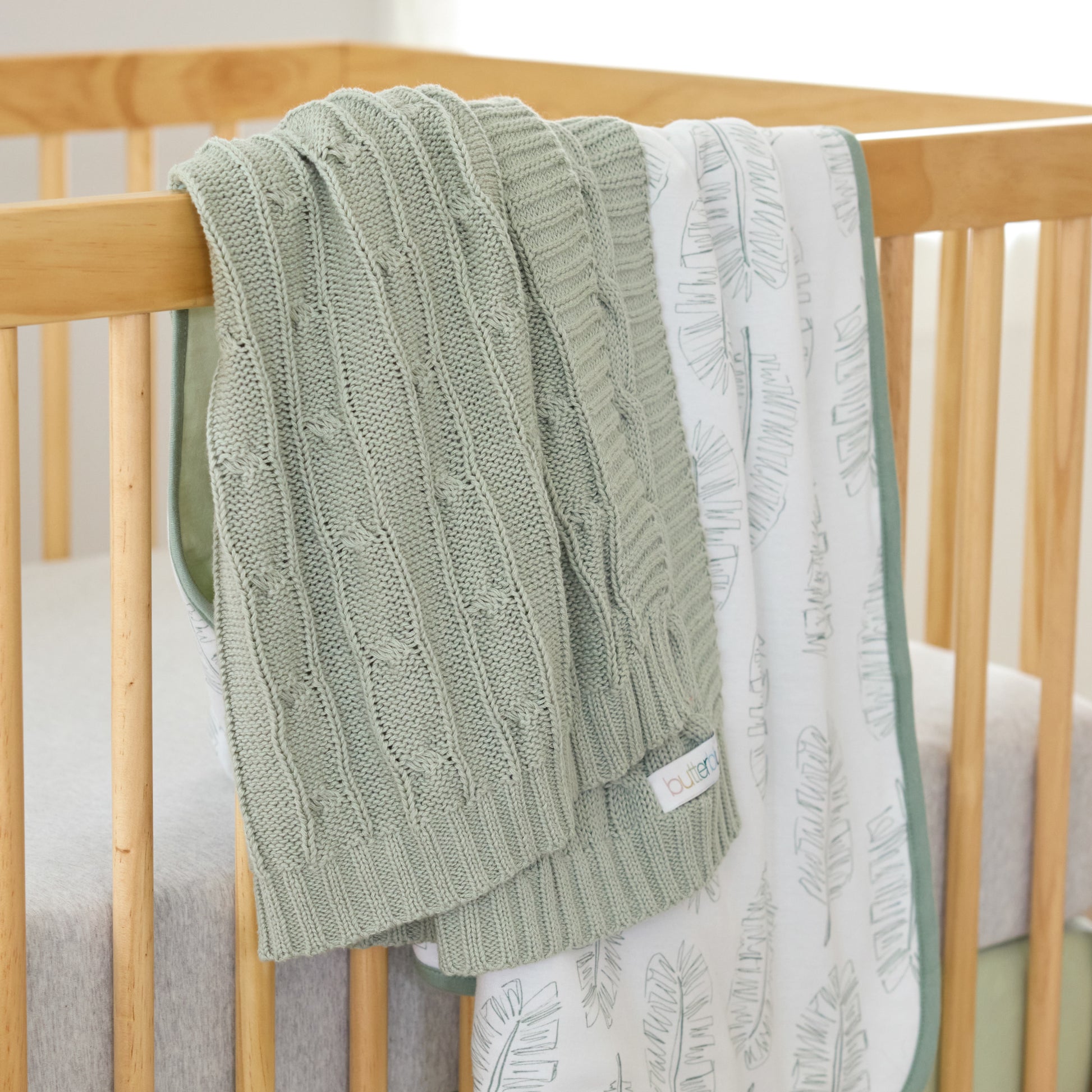 Green knitted blanket draped over a wooden crib with a white textured blanket underneath.
