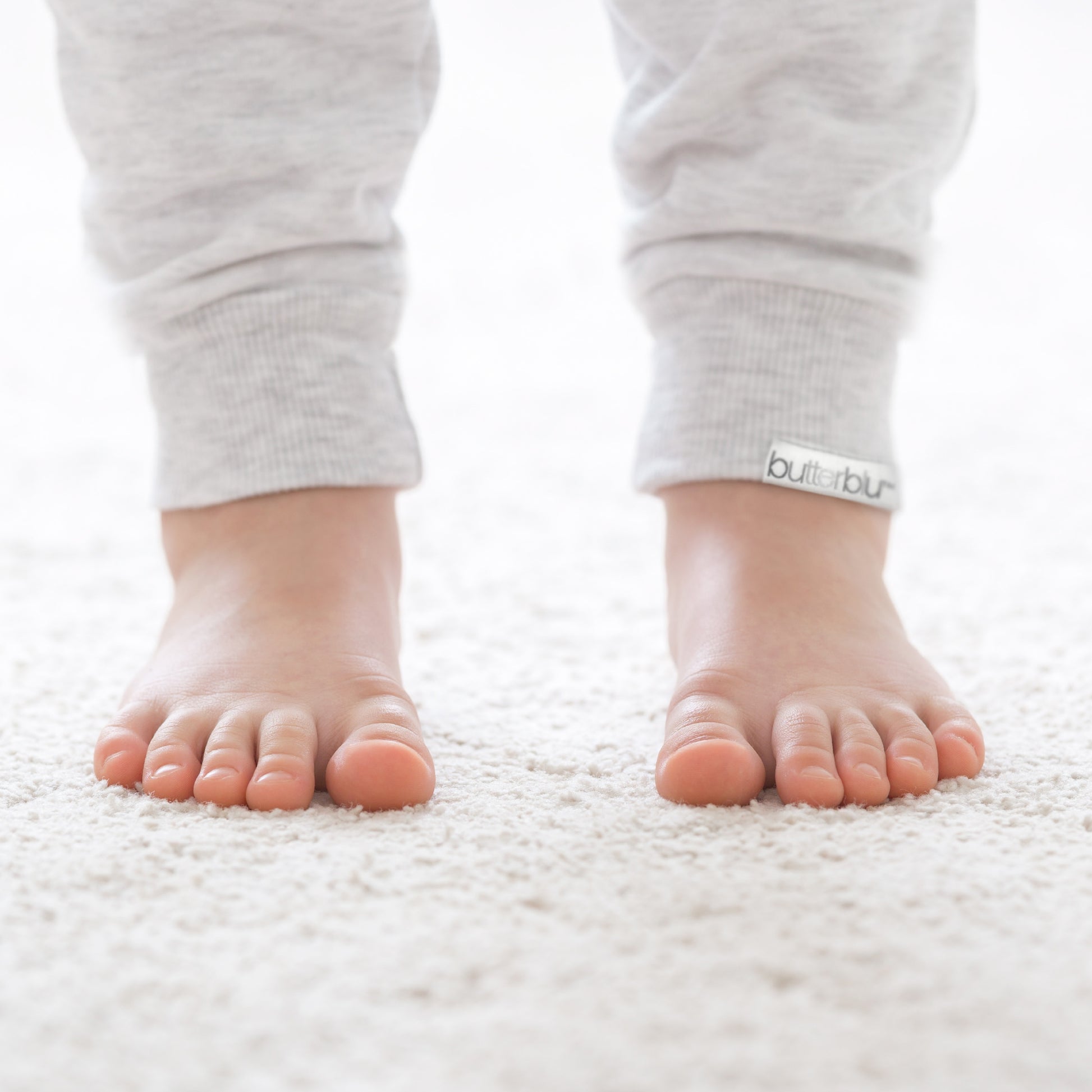 Baby's feet wearing gray pants with 'butterblu' brand logo on a white background